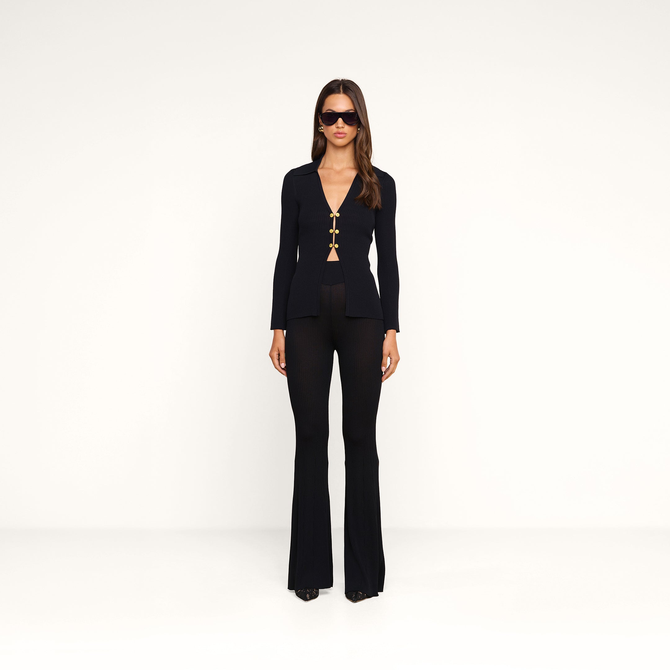 A woman stands against a plain white background wearing the fitted black Kaye Cardigan Top with gold clasps, paired with black flared pants, black shoes, and sunglasses. She looks straight ahead with her arms relaxed at her sides, creating a sleek travel set look.