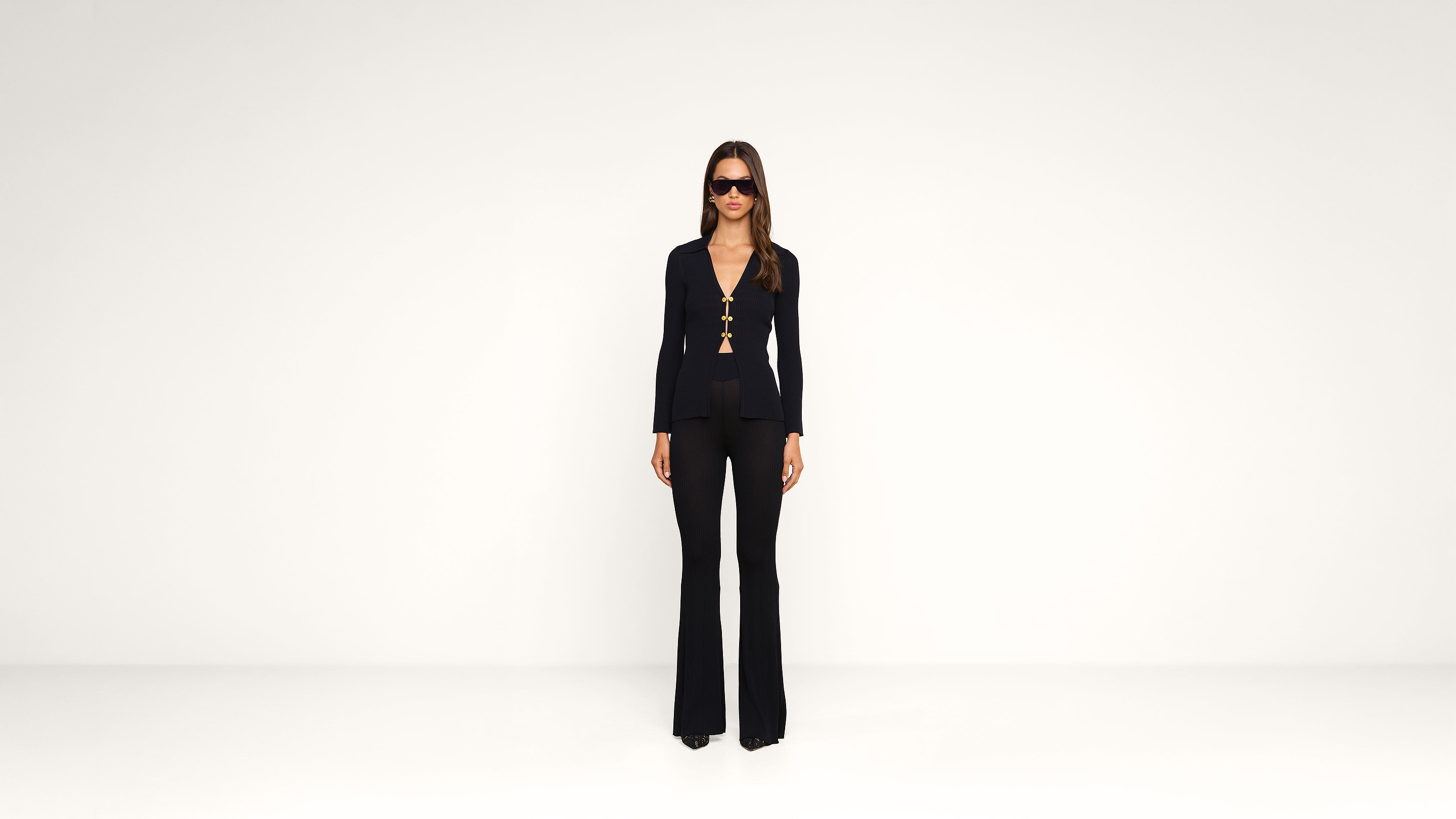 A woman stands against a plain white background wearing sunglasses, a black top with deep neckline and gold buttons, and black flared pants.