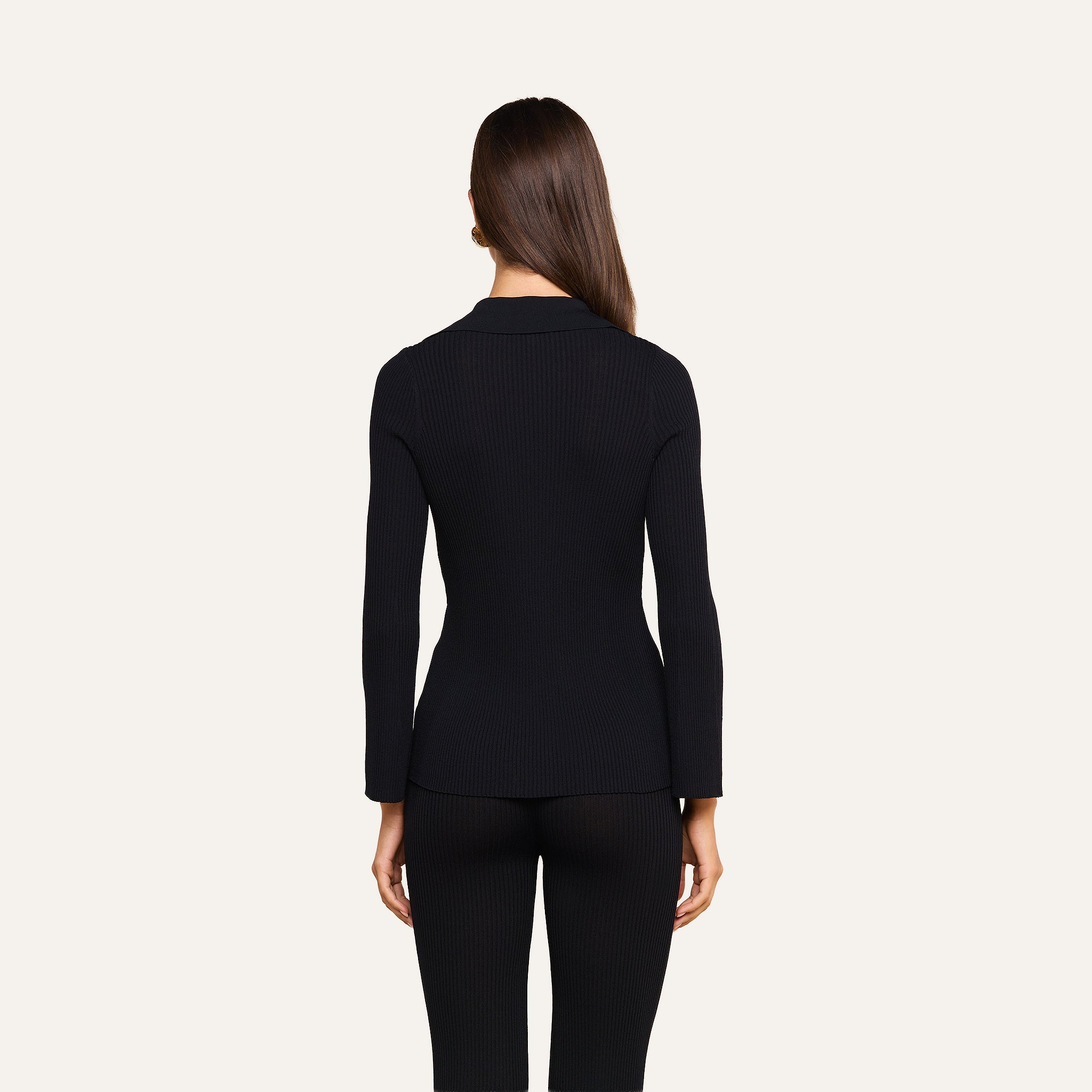 A woman with straight brown hair stands facing away, wearing the fitted black Kaye Cardigan Top and black pants, against a plain light background.
