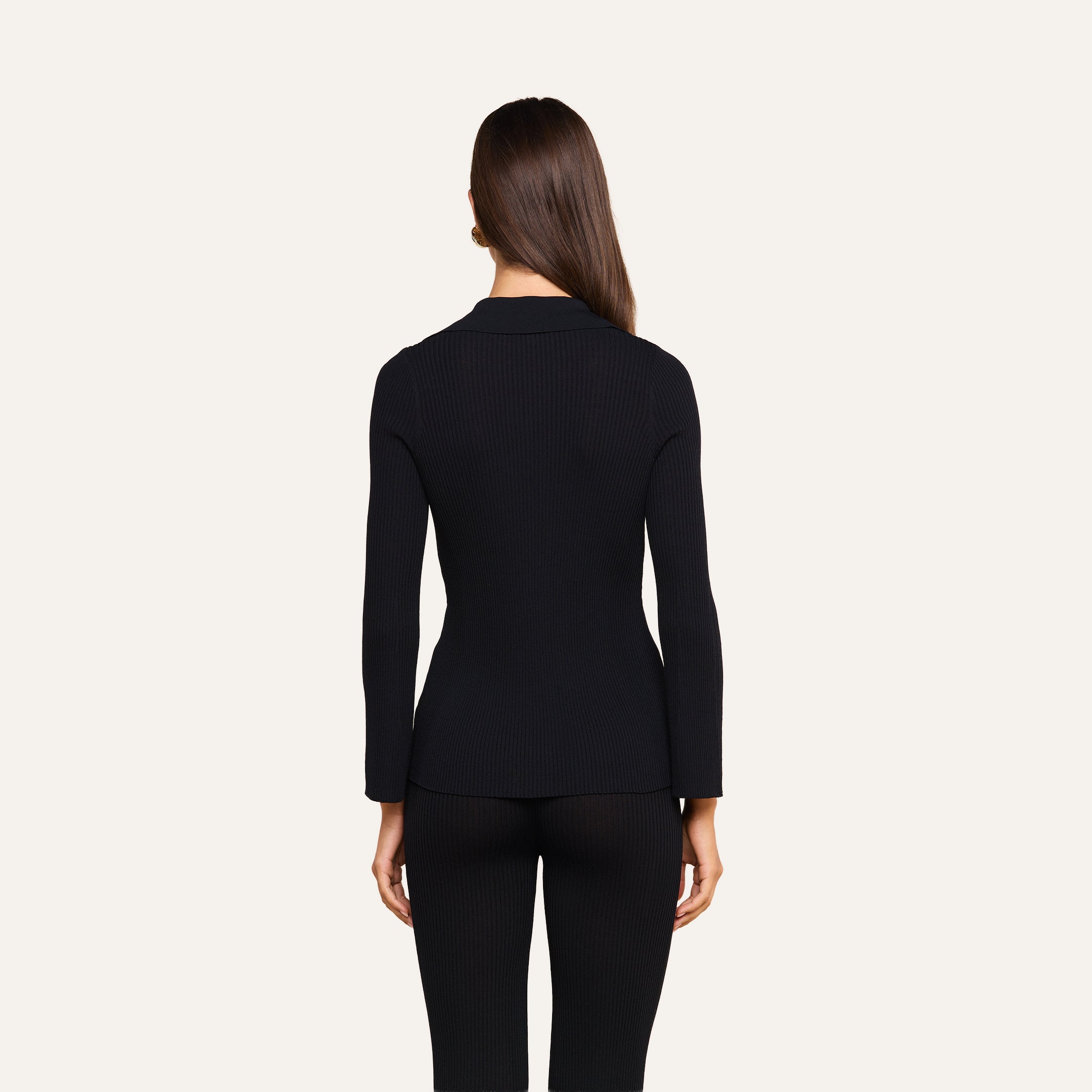 A woman with straight brown hair stands facing away, wearing the fitted black Kaye Cardigan Top and black pants, against a plain light background.