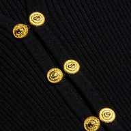 Six gleaming gold buttons adorn the ribbed black knit of the Kaye Cardigan Top, arranged in two asymmetrical columns. The rich texture of the fabric and striking metallic buttons are clearly showcased, offering an elegant look that pairs effortlessly with any travel set.