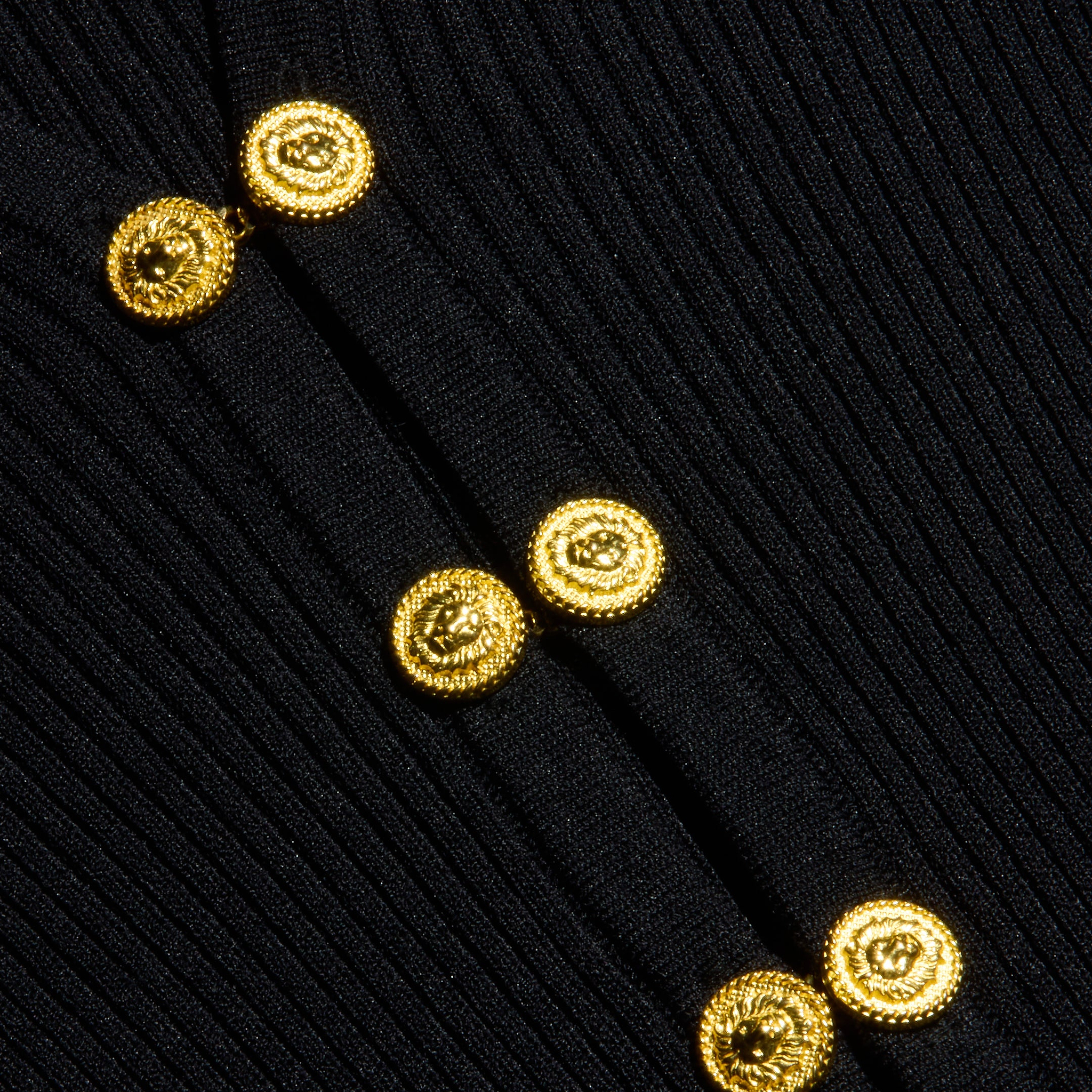 Six gleaming gold buttons adorn the ribbed black knit of the Kaye Cardigan Top, arranged in two asymmetrical columns. The rich texture of the fabric and striking metallic buttons are clearly showcased, offering an elegant look that pairs effortlessly with any travel set.