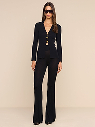 A woman stands against a white background wearing the Conan Knit Pant, a pair of fitted black high-rise flared pants with gold buttons. She pairs them with black heels and dark sunglasses, her long hair loose as she looks straight at the camera.