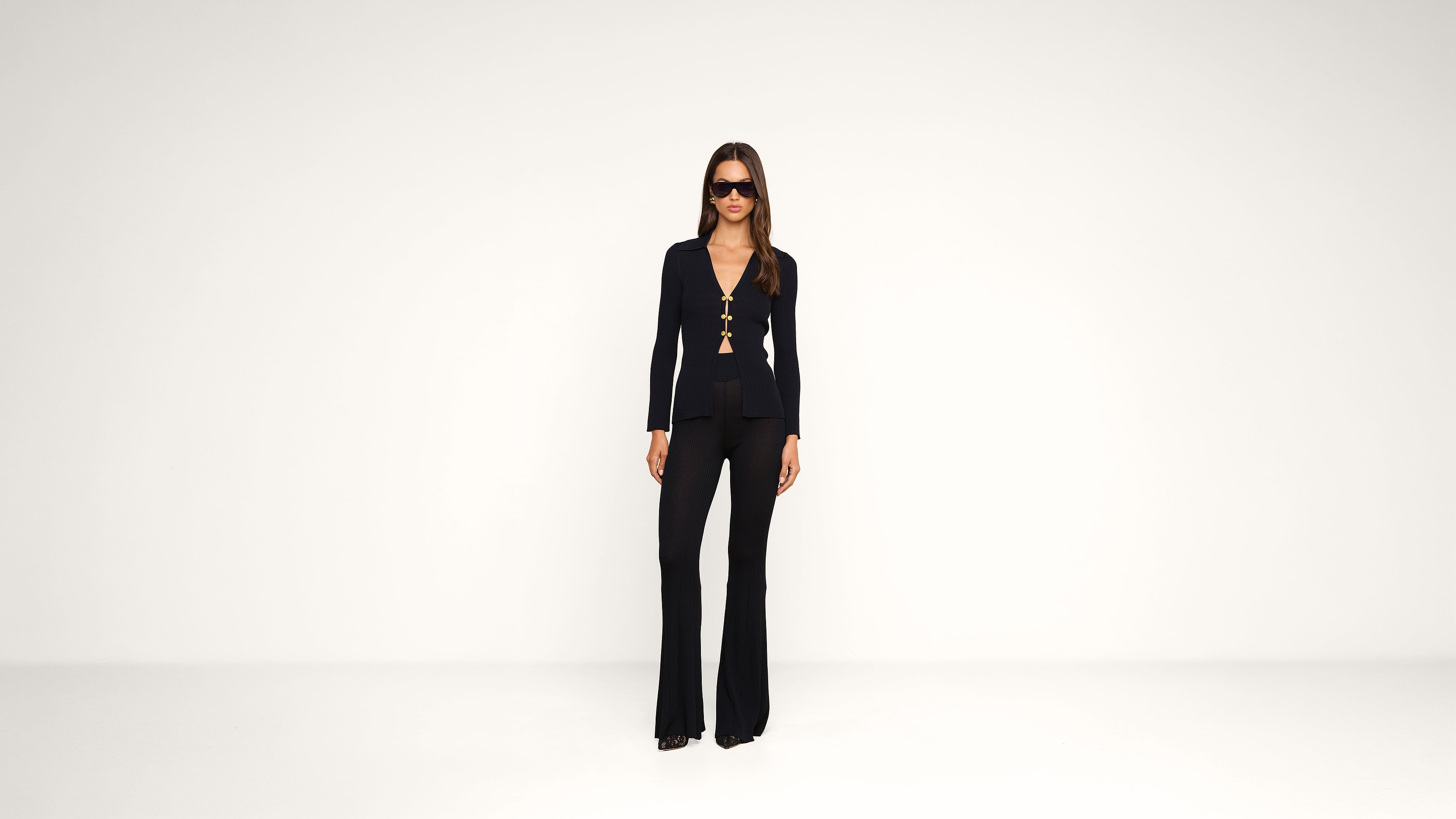 A person stands in a white room wearing black flared pants, a fitted black top with gold accents, black sunglasses, and heeled shoes.
