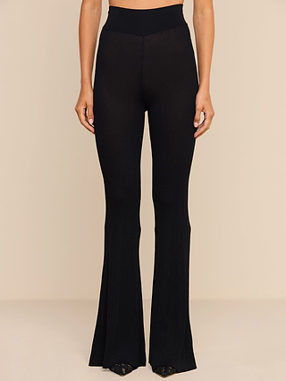 A person is shown from the waist down wearing the Conan Knit Pant, a high rise, black ribbed-knit flared pant, standing against a beige background.