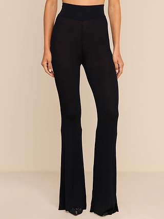 A person wearing the Conan Knit Pant in black with a high-rise, flared silhouette and pointed shoes, standing against a plain beige background.