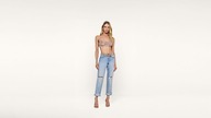 Woman standing against a plain white background, wearing a patterned crop top, light blue ripped jeans, and beige high-heeled sandals.