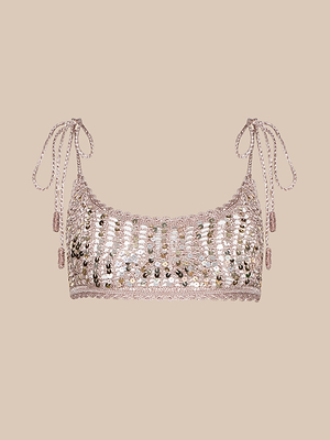 The Taline Crocheted Bralette is a sequined summer top featuring thin braided straps tied in bows, with light pink and metallic accents that shine against a plain beige background.