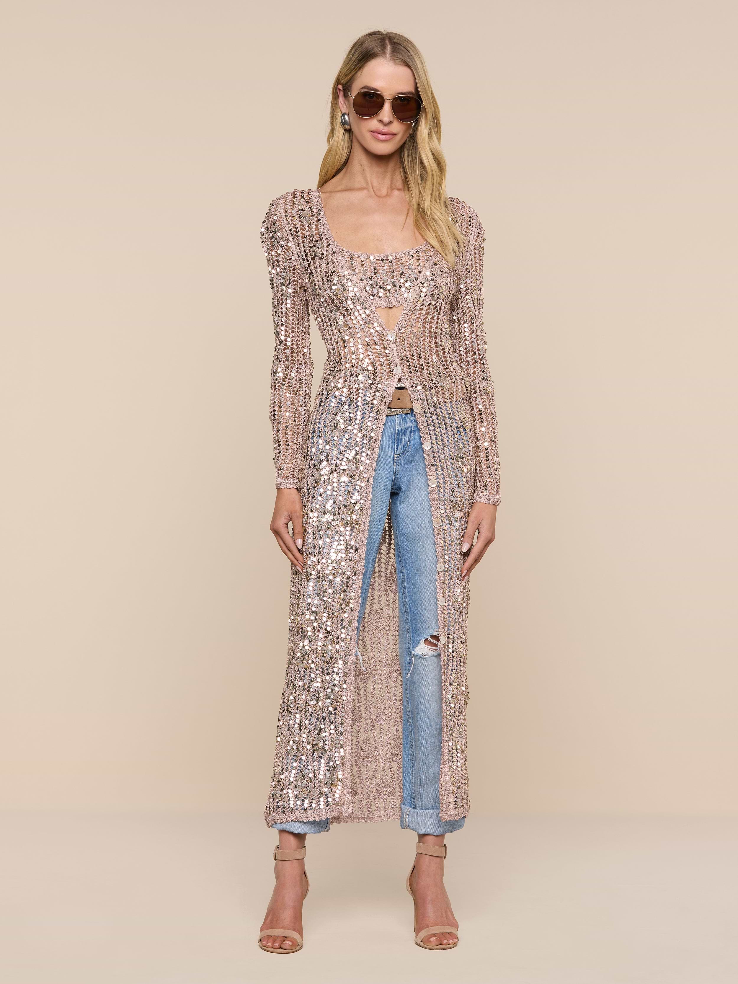 A woman wearing sunglasses, the Sanya Crocheted Cardigan—a long, sequined crochet cardigan—over a matching top, paired with light blue ripped jeans and nude heels, stands against a plain white background. The Sanya Crocheted Cardigan is perfect as a stylish swimsuit cover-up or vacation outfit.