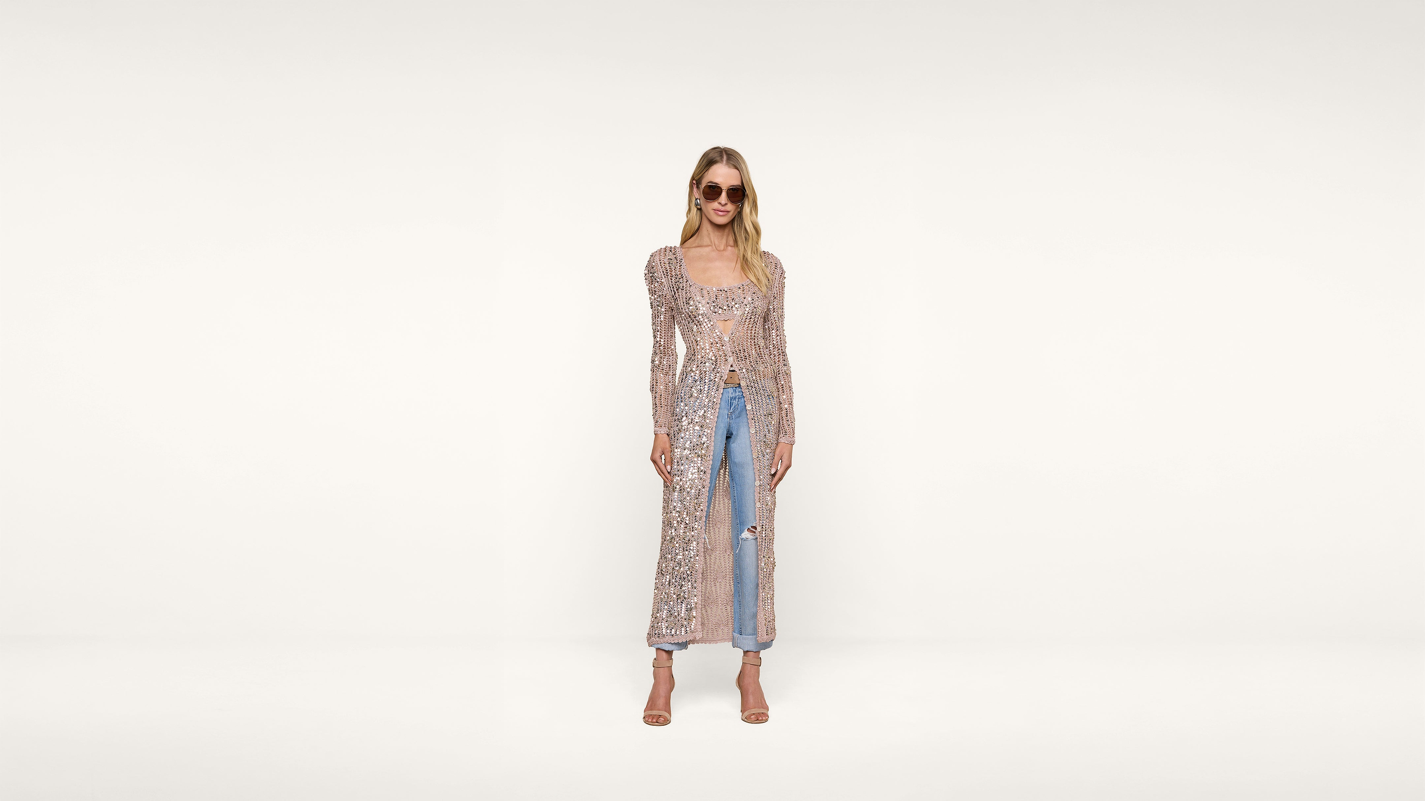 Woman wearing sunglasses, a long sheer floral-patterned duster, light-wash ripped jeans, and nude heels stands against a plain white background, smiling with arms relaxed at her sides.