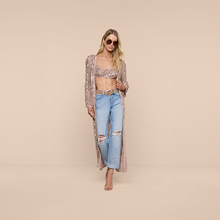 A woman wearing sunglasses, a sequined crochet crop top, the Sanya Crocheted Cardigan layered over it, ripped light blue jeans, and nude heels poses confidently against a plain beige background—an effortlessly chic look perfect for your next summer vacation.