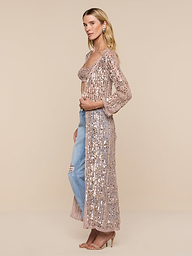 A woman stands sideways against a plain white background, wearing the Sanya Crocheted Cardigan in long, light pink sequined crochet over a beige top and ripped blue jeans, paired with nude heels. This versatile look makes for a stylish vacation outfit or a glamorous swimsuit cover up.