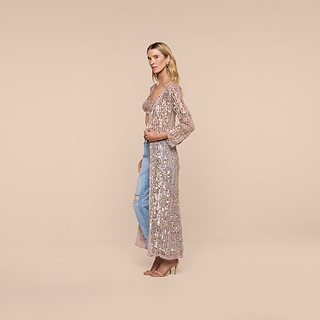 A woman stands sideways against a beige background, wearing the Sanya Crocheted Cardigan as a stylish sheer cover-up over a pink top and ripped blue jeans, paired with beige high heels.