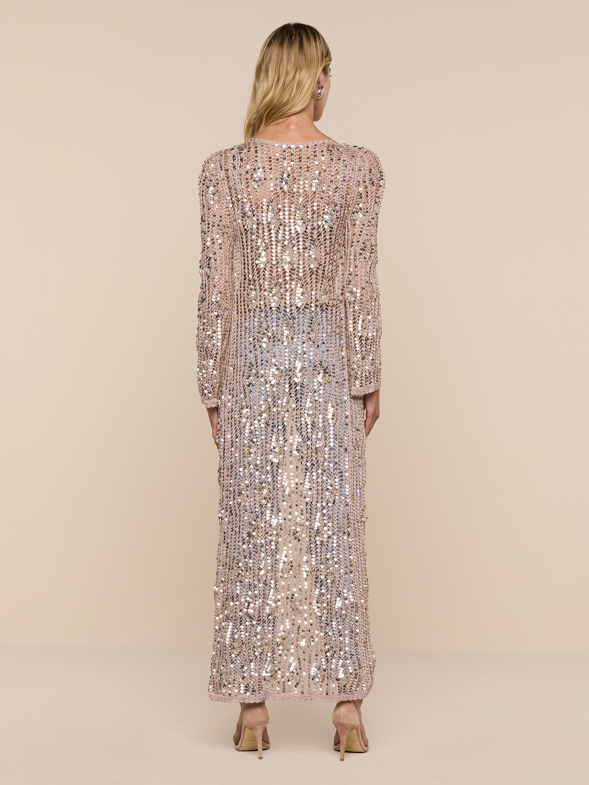 A person with long blonde hair stands facing away, wearing the Sanya Crocheted Cardigan and heeled shoes. The cardigan is sparkling, long-sleeved, and floor-length. The background is plain and white.