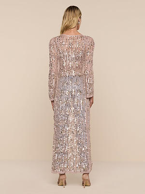 A woman with long blonde hair faces away in the Sanya Crocheted Cardigan, a sheer nude ankle-length cover-up adorned with silver sequins and paired with nude heels—ideal for adding glamour to any swimsuit look against a beige backdrop.