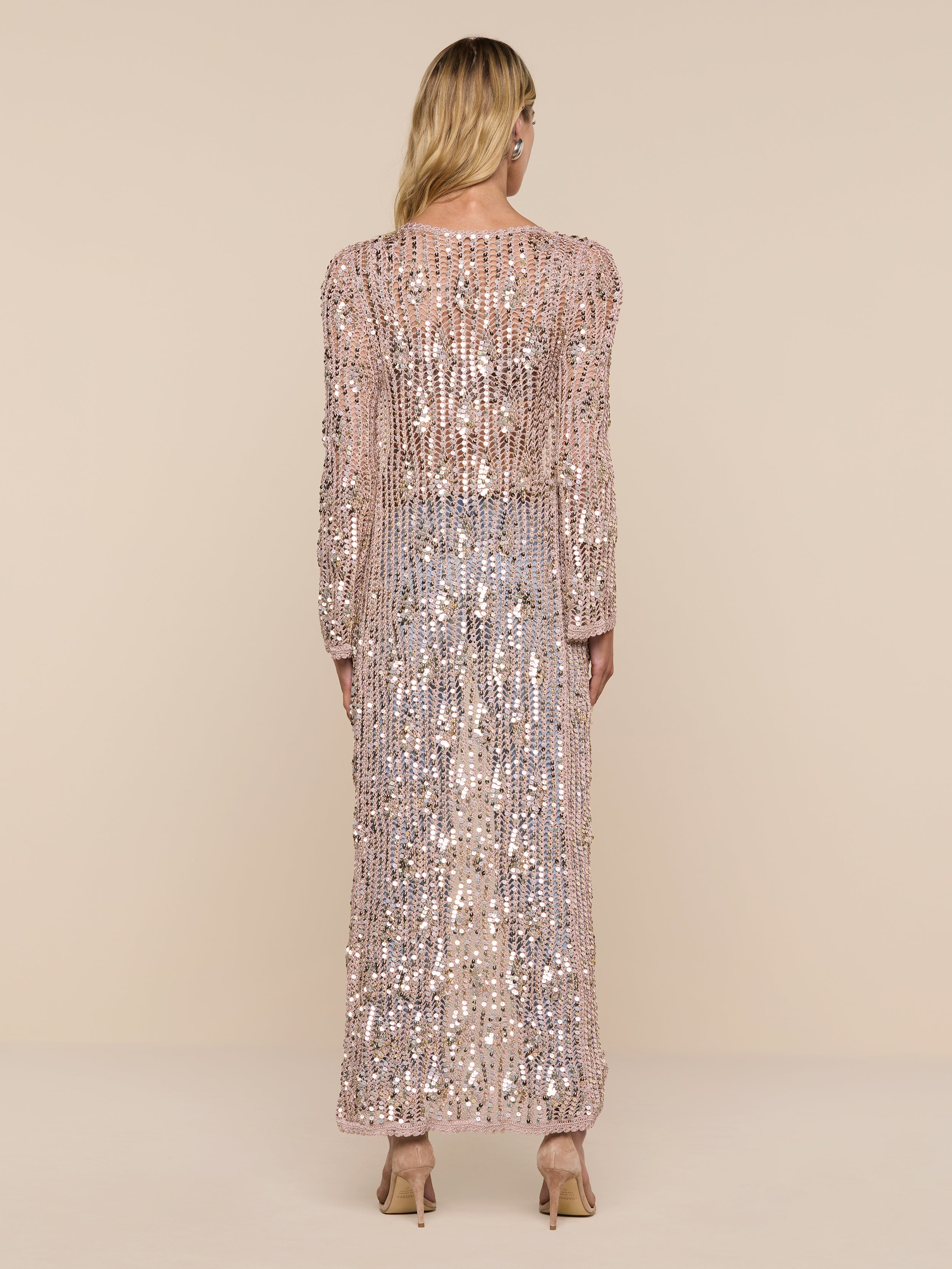 A person with long blonde hair stands facing away, wearing the Sanya Crocheted Cardigan and heeled shoes. The cardigan is sparkling, long-sleeved, and floor-length. The background is plain and white.