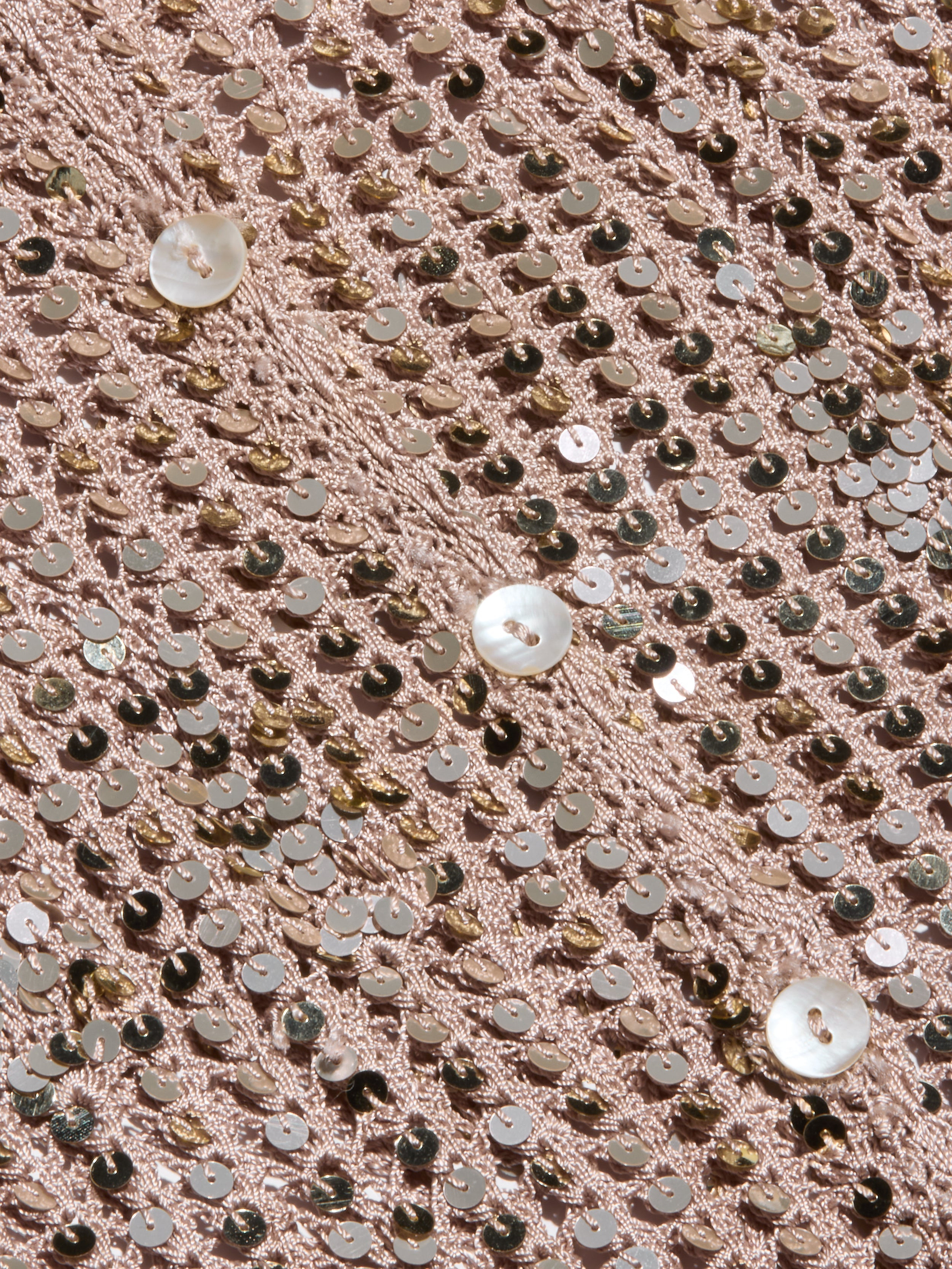 Close-up of shiny, rose gold sequins sewn onto fabric in a diagonal pattern, reminiscent of the Sanya Crocheted Cardigan, with a few larger, round, white buttons scattered on top—perfect for creating a standout vacation outfit.