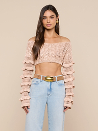 A woman wearing the Fey Crocheted Top, a beige off-shoulder piece showcasing handmade artistry with tiered sleeves, paired with light blue high-waisted jeans and standing against a plain light background.