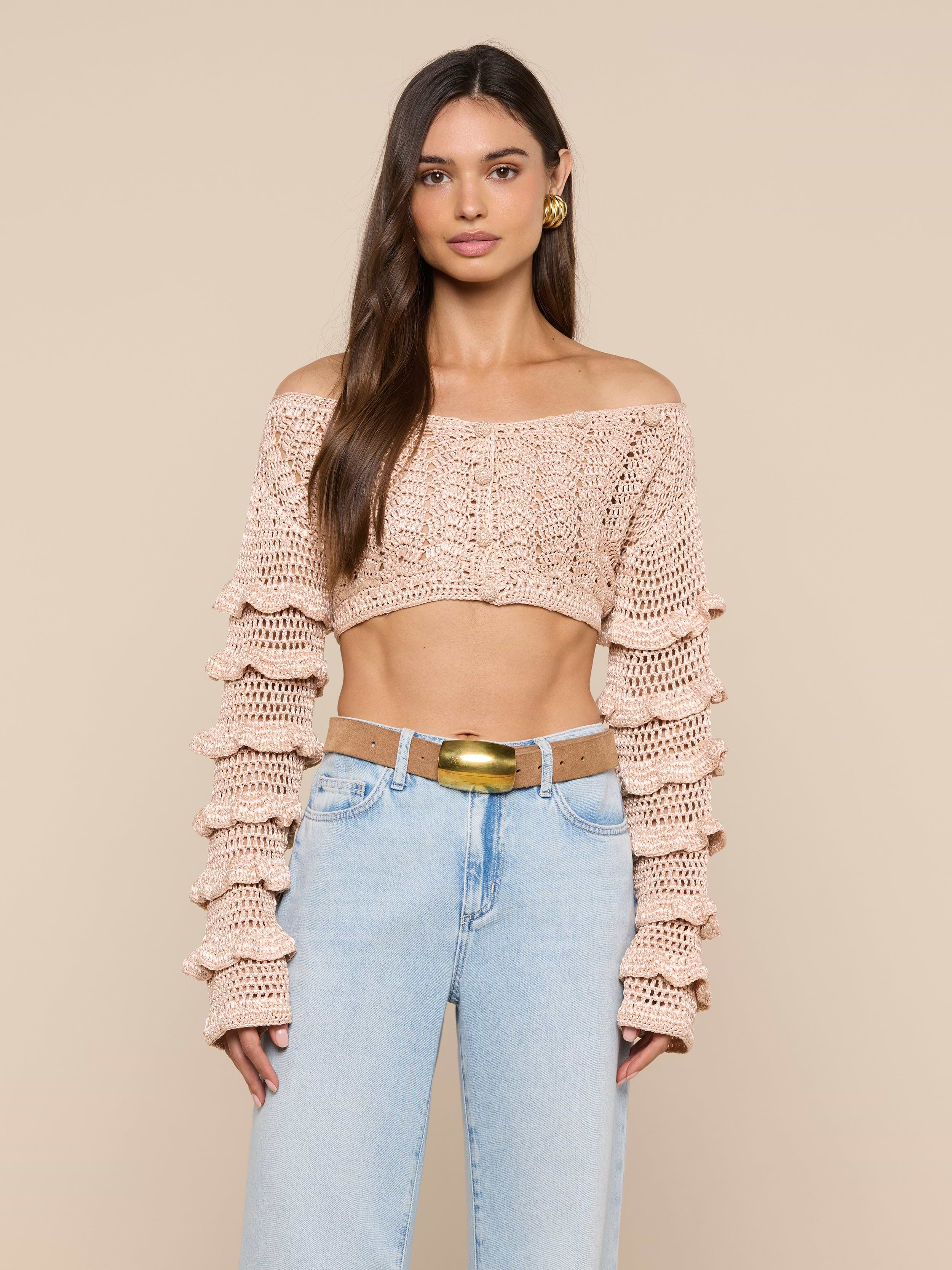 A woman wearing the Fey Crocheted Top, a beige off-shoulder piece showcasing handmade artistry with tiered sleeves, paired with light blue high-waisted jeans and standing against a plain light background.