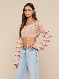 A woman wearing the Fey Crocheted Top—a beige, off-shoulder design with ruffled sleeves—paired with high-waisted light blue jeans, stands against a plain background. The piece showcases handmade artistry in its beautifully knit details.