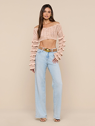 A woman stands against a plain white background, wearing the Fey Crocheted Top—a beige off-shoulder piece with ruffled detailing—paired with light blue high-waisted wide-leg jeans, brown sandals, and a gold-buckled belt. The look accentuates the handmade artistry of the Fey Crocheted Top’s delicate design.