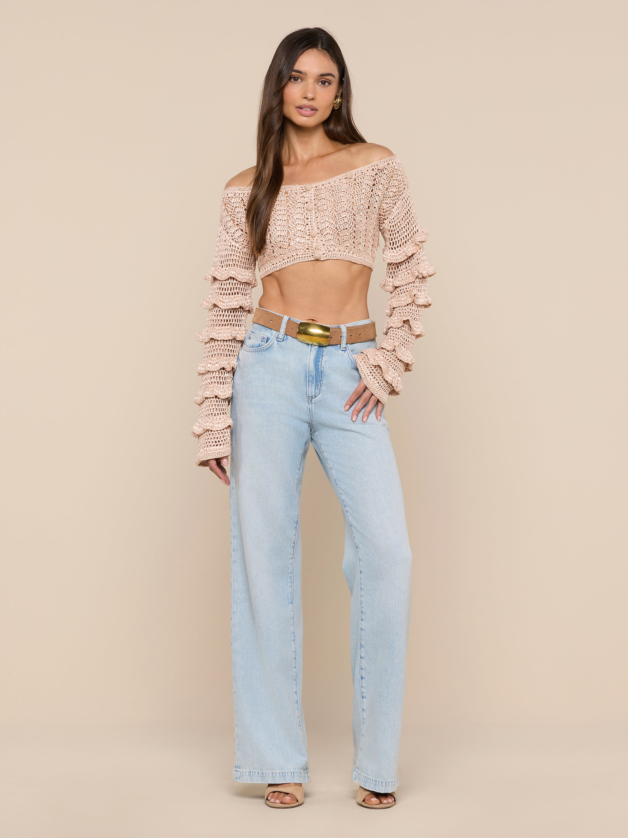 A woman stands against a plain white background, wearing the Fey Crocheted Top—a beige off-shoulder piece with ruffled detailing—paired with light blue high-waisted wide-leg jeans, brown sandals, and a gold-buckled belt. The look accentuates the handmade artistry of the Fey Crocheted Top’s delicate design.