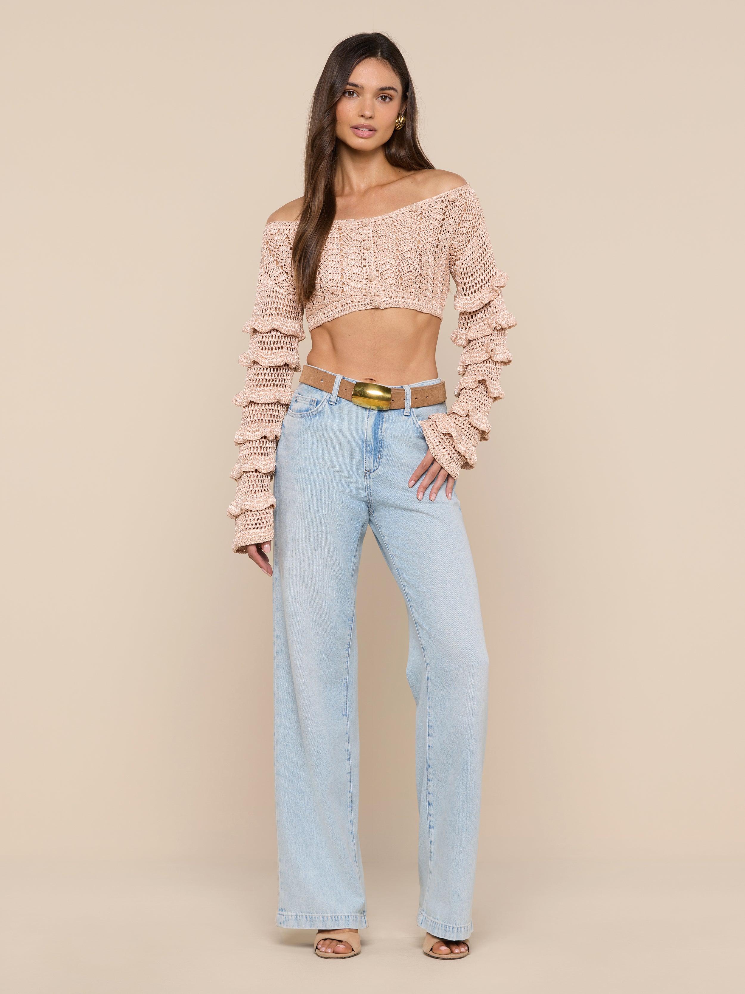 A woman stands against a plain white background, wearing the Fey Crocheted Top—a beige off-shoulder piece with ruffled detailing—paired with light blue high-waisted wide-leg jeans, brown sandals, and a gold-buckled belt. The look accentuates the handmade artistry of the Fey Crocheted Top’s delicate design.