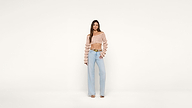 A woman stands against a plain white background wearing a pink off-the-shoulder ruffle top, light blue high-waisted wide-leg jeans, and heeled sandals. She poses with one hand on her hip.