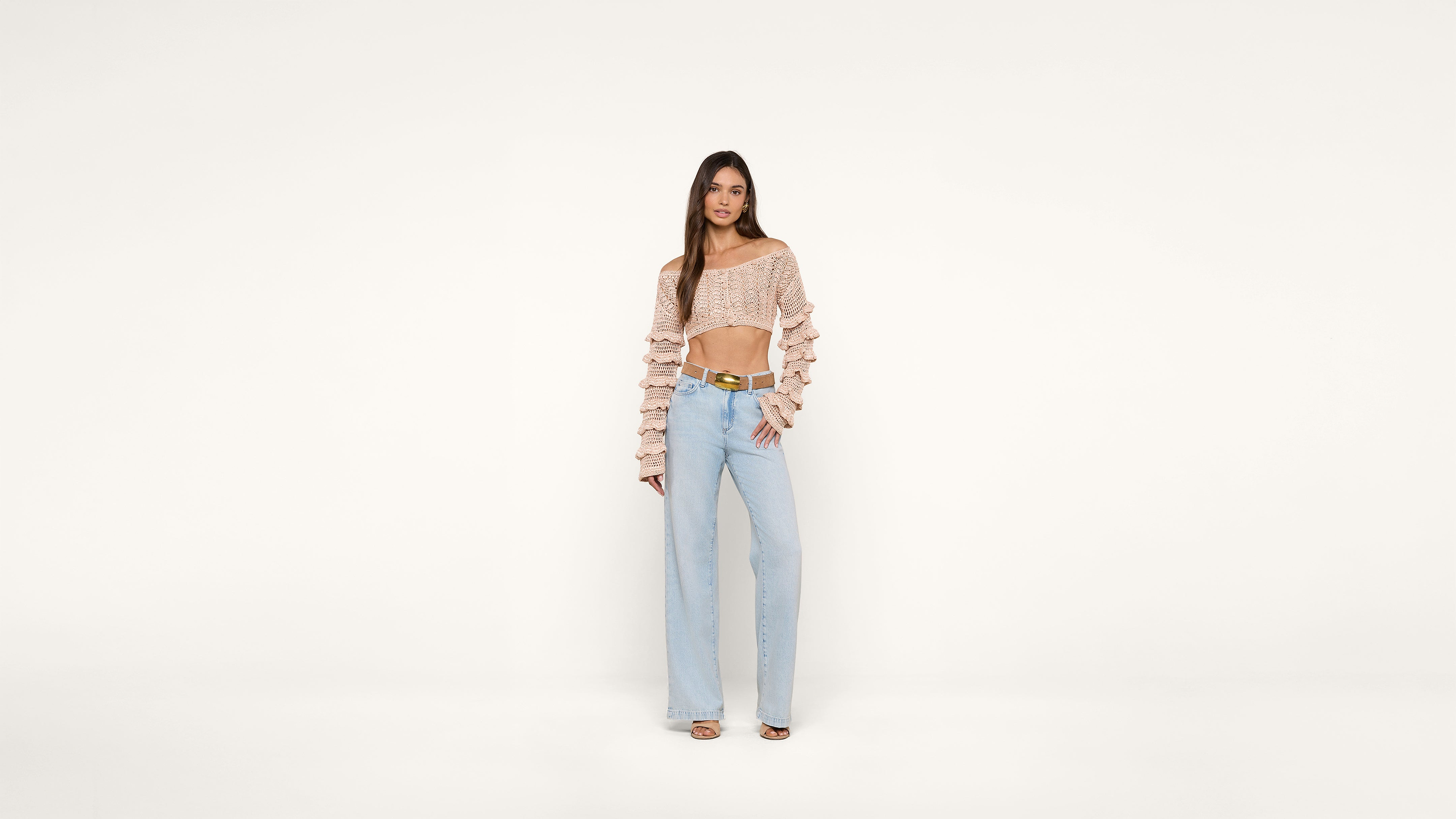 A woman stands against a plain white background wearing a pink off-the-shoulder ruffle top, light blue high-waisted wide-leg jeans, and heeled sandals. She poses with one hand on her hip.