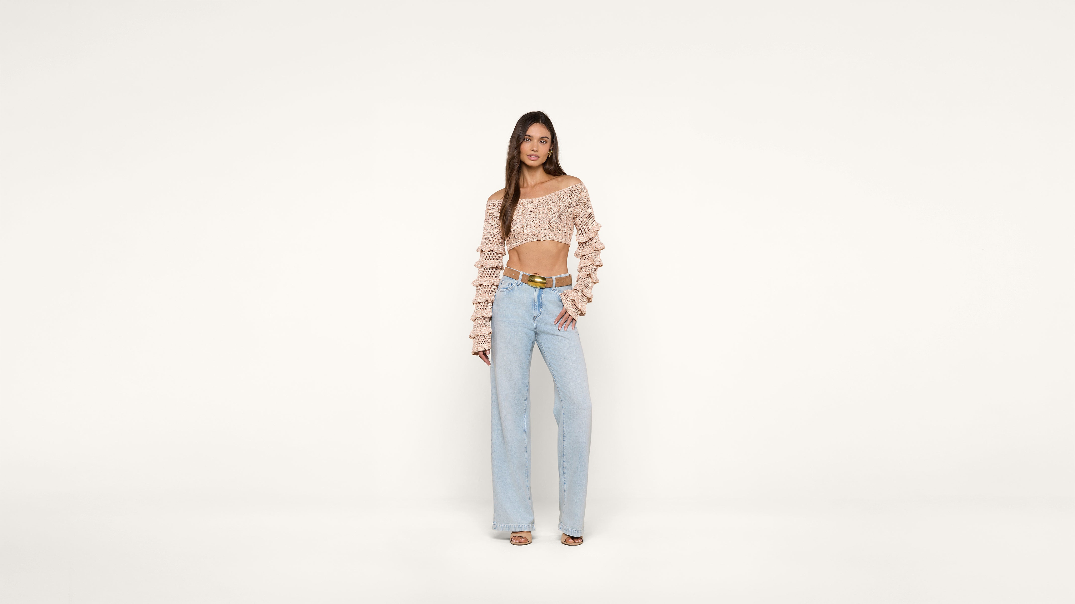 A woman stands against a plain white background wearing a pink off-the-shoulder ruffle top, light blue high-waisted wide-leg jeans, and heeled sandals. She poses with one hand on her hip.