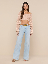 A woman stands facing forward, showcasing the Fey Crocheted Top—an artistry-inspired, handmade cropped off-shoulder knit with ruffled sleeves—paired with light blue high-waisted wide-leg jeans, a gold belt, and open-toe heels against a plain background.