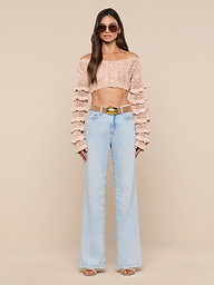 A woman wearing sunglasses, the Fey Crocheted Top—a cropped knit sweater with ruffled sleeves that showcases handmade artistry—paired with light blue high-waisted flared jeans, open-toe heels, and a gold belt, standing against a plain white background.