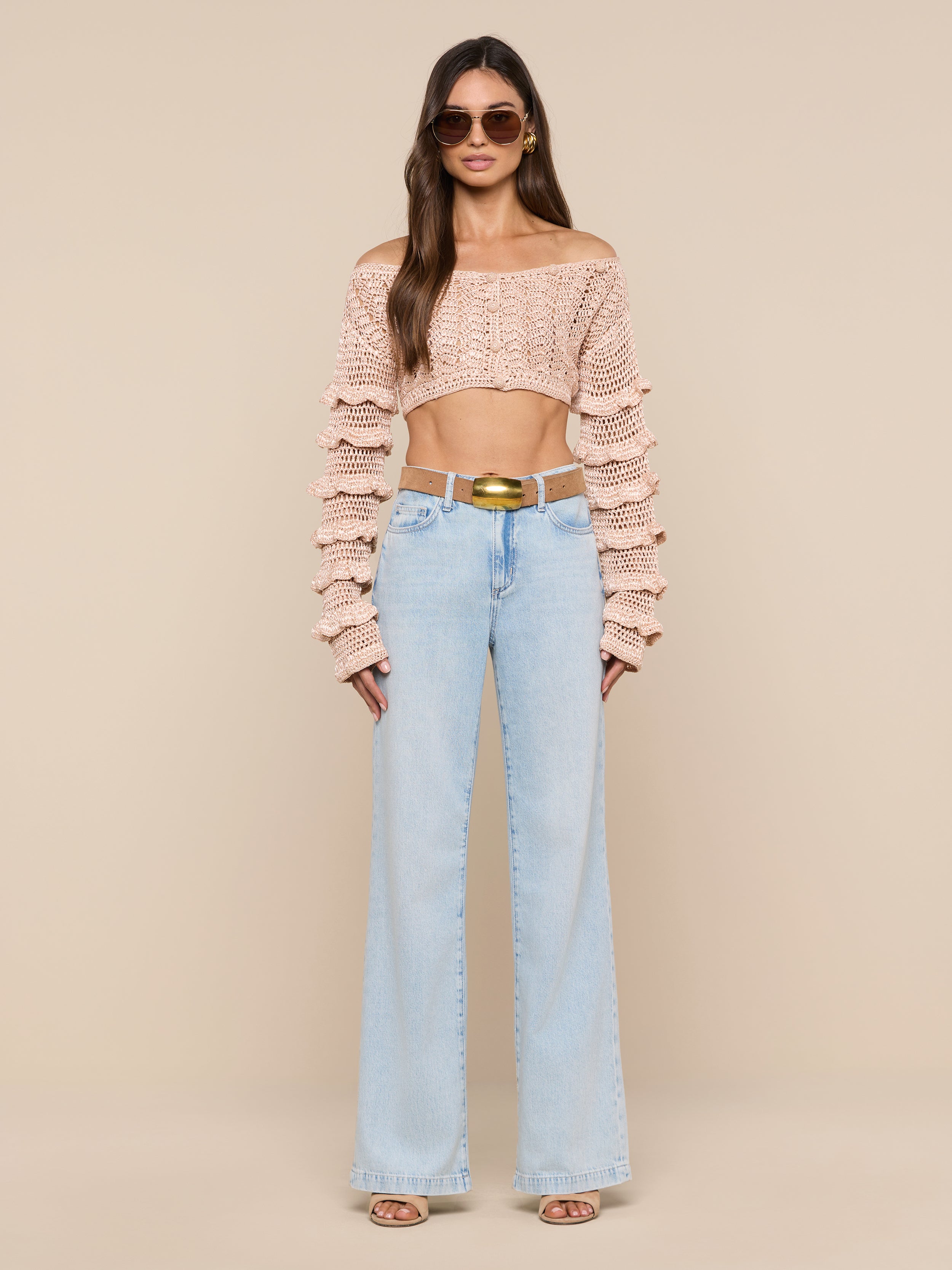 A woman wearing sunglasses, the Fey Crocheted Top—a cropped knit sweater with ruffled sleeves that showcases handmade artistry—paired with light blue high-waisted flared jeans, open-toe heels, and a gold belt, standing against a plain white background.