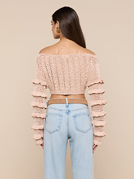 A person stands facing away, wearing light blue jeans and the Fey Crocheted Top—a pink off-shoulder design with ruffled sleeves—showcasing handmade artistry against a plain, light background.