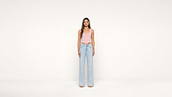A woman stands facing forward against a plain white background, wearing a light pink sleeveless top and high-waisted, wide-leg light blue jeans with open-toe shoes.