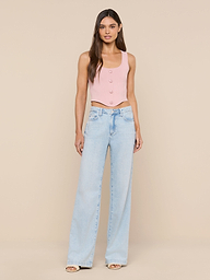 A woman stands facing forward, wearing the Dove Knit Corset Top—a pink fitted sleeveless piece with a curved hemline—paired with light blue wide-leg jeans. She has long, straight hair and is posed against a plain white background.