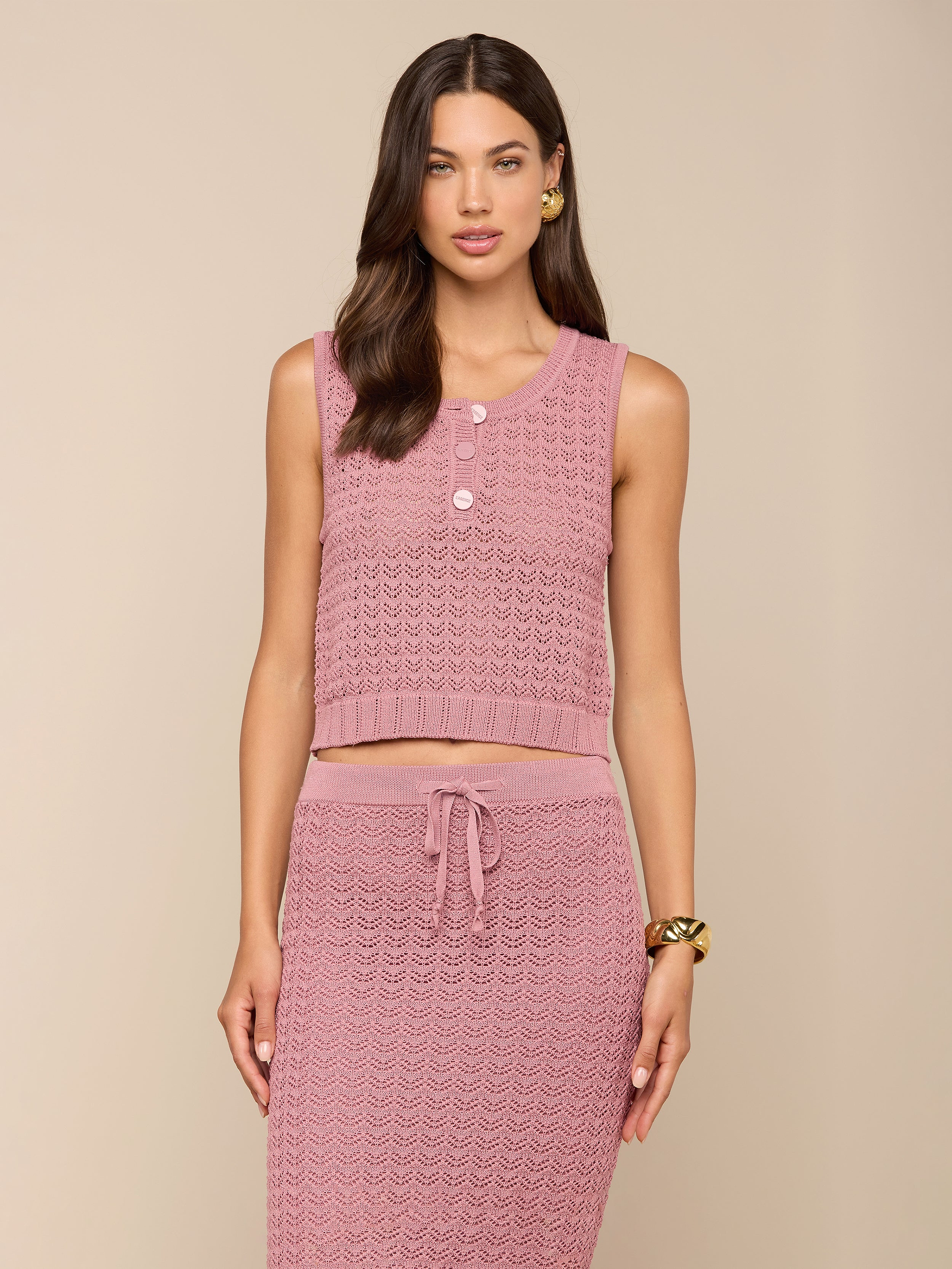 Una donna indossa la canottiera Carven Pointelle Knit Tank in rosa testurizzato con bottoni, abbinata a una gonna a vita alta con coulisse e accessori dorati, su uno sfondo beige.