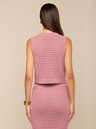 Una donna con lunghi capelli castani indossa una canottiera Carven Pointelle Knit e una gonna abbinata, in piedi con le spalle allobiettivo su uno sfondo beige. Al polso destro indossa un braccialetto doro.