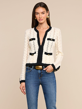 A woman with long brown hair wears the Zephyra Pointelle Knit Cardigan in cream lace with black trim and gold buttons, paired with blue jeans, a wide black belt featuring a gold buckle, and gold earrings, standing against a beige background.