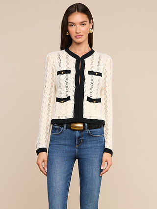 A woman with long brown hair wears the Zephyra Pointelle Knit Cardigan—a cozy cream knit featuring black trim and buttons—paired with blue jeans, a black belt with a gold buckle, and gold earrings, standing against a beige background.