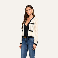 A woman wearing the cream Zephyra Pointelle Knit Cardigan with black trim and decorative buttons, paired with a black top, blue jeans, and gold earrings, stands against a plain light background.