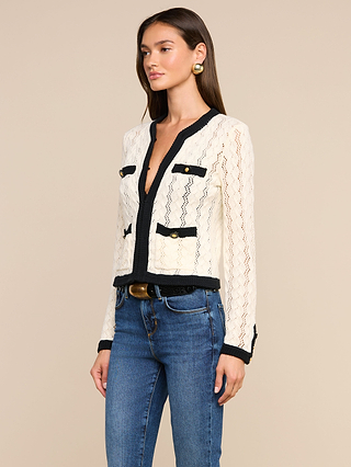 A woman wearing the Zephyra Pointelle Knit Cardigan—a cozy cream lace pointelle knit featuring black trim and gold buttons—paired with blue jeans, stands against a beige background.