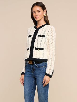 A woman wearing the Zephyra Pointelle Knit Cardigan, a cream lace jacket with black trim and gold buttons reminiscent of a cozy cardigan, paired with blue jeans and a black belt, stands against a plain beige background.
