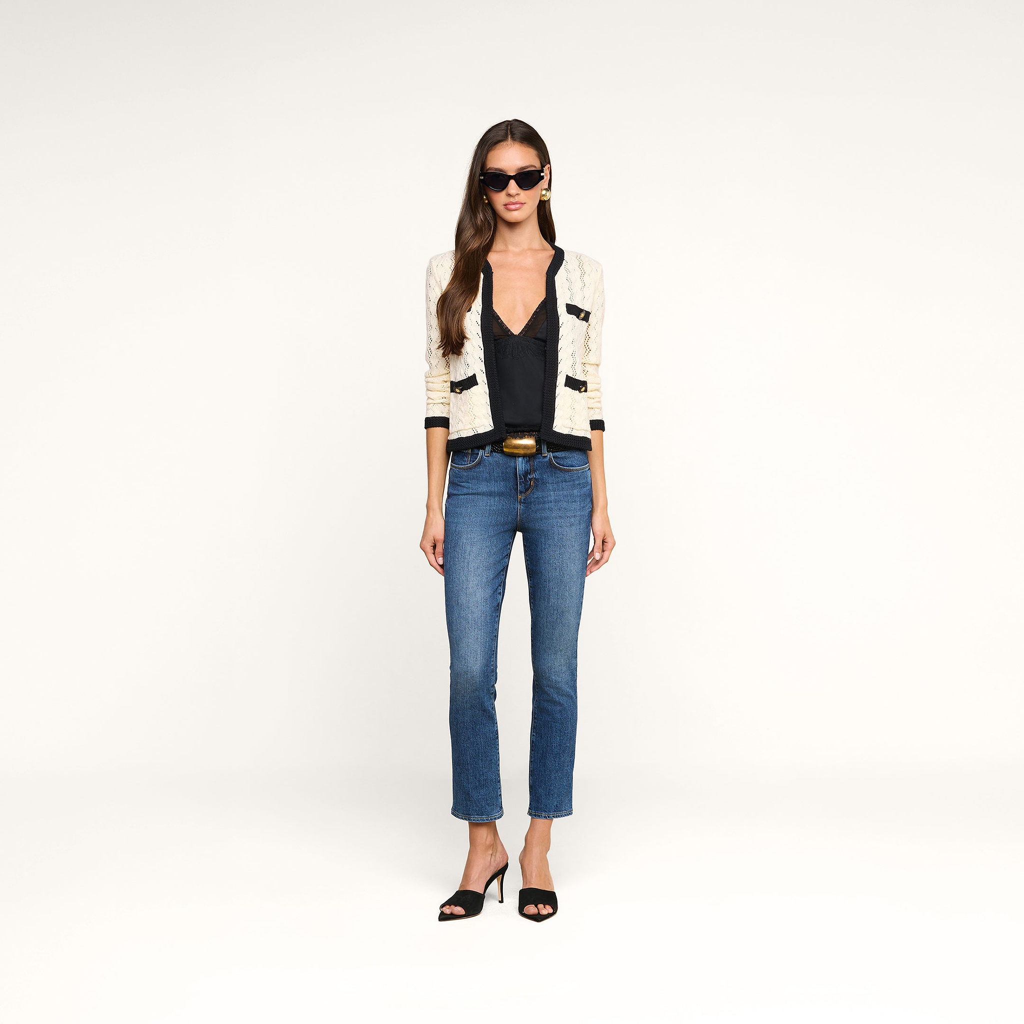 A woman wearing sunglasses, a cream-colored Zephyra Pointelle Knit Cardigan with decorative buttons, a black top, blue jeans, and black heeled sandals stands against a plain white background.