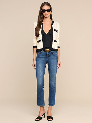 A woman wearing sunglasses, a cozy cream and black Zephyra Pointelle Knit Cardigan, black top, blue jeans, a belt, and black-heeled sandals stands facing forward against a plain beige background.