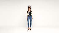 Woman in sunglasses wearing a cream cardigan with black trim, black top, blue jeans, and black heels stands against a plain white background.