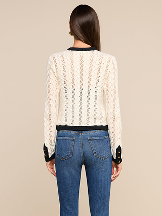 A woman with straight brown hair is shown from the back, wearing the Zephyra Pointelle Knit Cardigan—a white lace long-sleeve top with black trim—paired with blue jeans. Her cozy cardigan style stands out against a plain beige background.