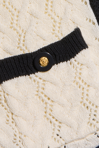 Close-up of a cream cable-knit fabric with a black ribbed trim, decorative gold and black button, and a small pocket—perfect details that define the cozy cardigan charm of the Zephyra Pointelle Knit Cardigan.