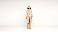 A woman stands in a beige outfit, including a long coat, wide-leg pants, and heeled shoes, against a plain white background.