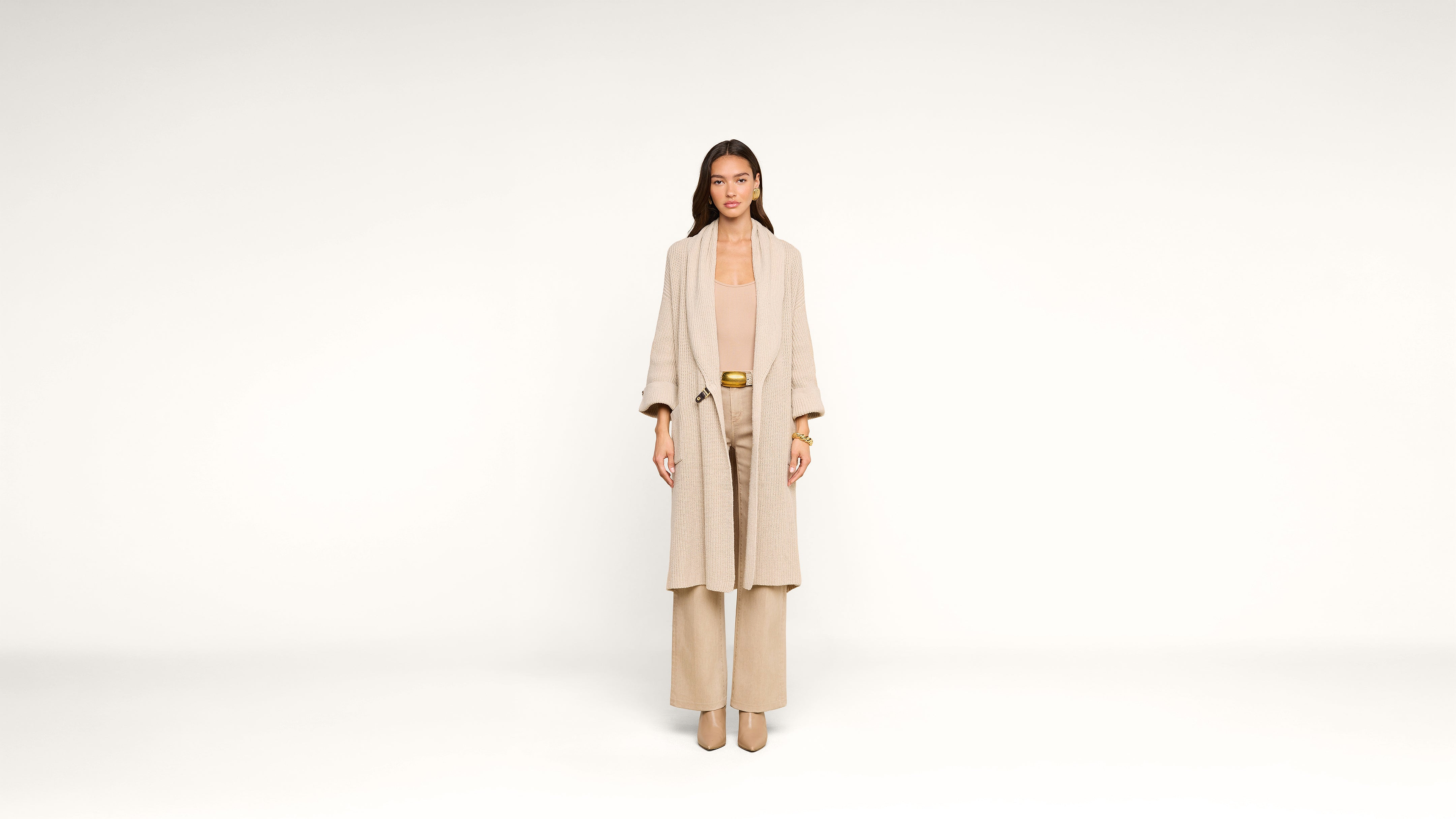 A woman stands in a beige outfit, including a long coat, wide-leg pants, and heeled shoes, against a plain white background.
